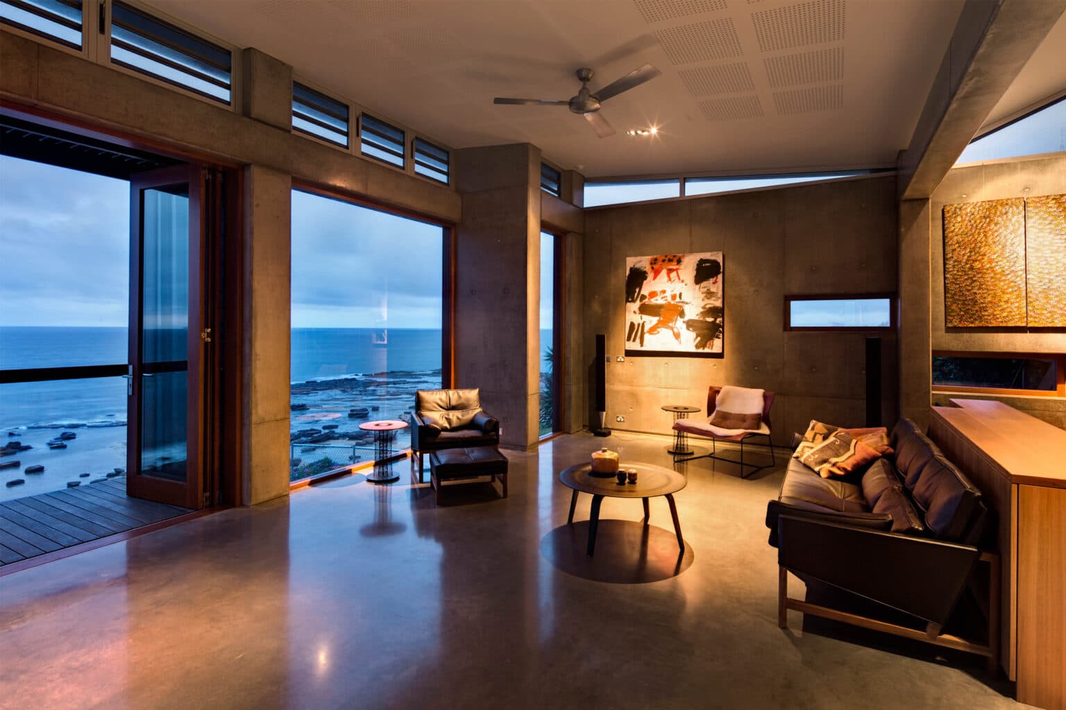 Modern living room with ocean view, abstract art, leather chairs, and warm lighting.