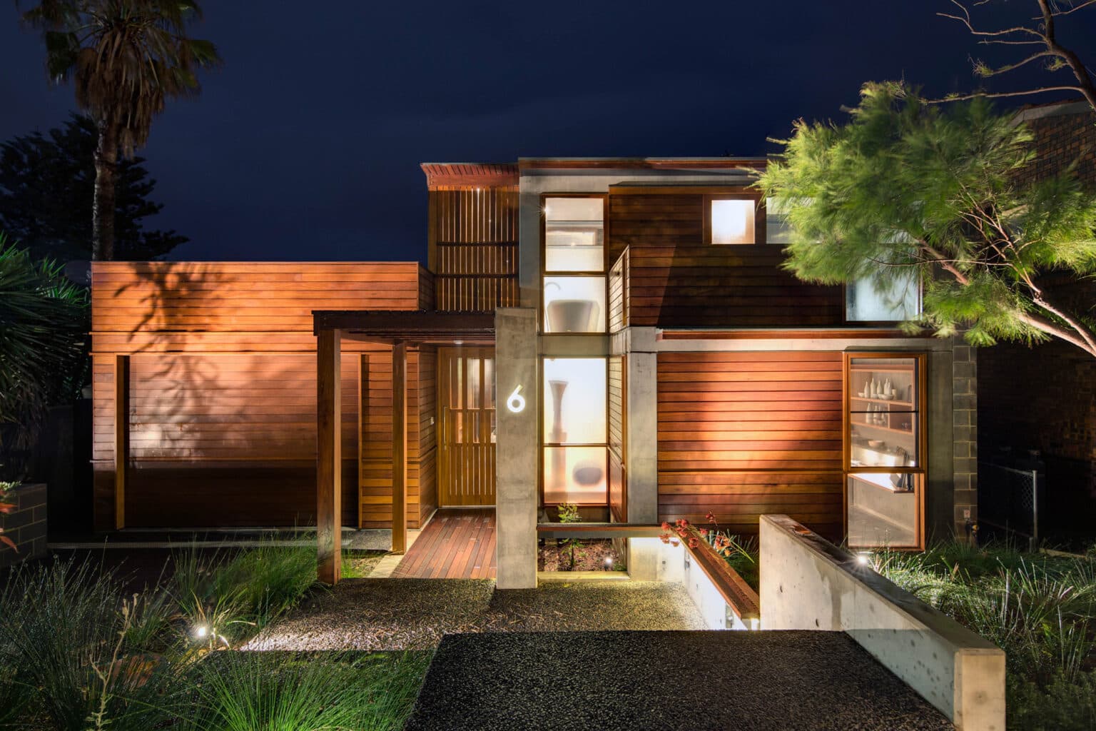 Modern wooden house with large windows, glowing facade, and landscaped garden illuminated at night.