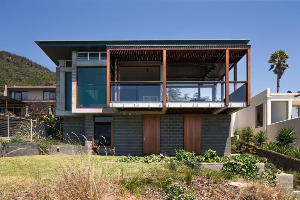 Modern two-story house with large glass windows, wooden accents, surrounded by greenery and palm trees under a clear blue ...