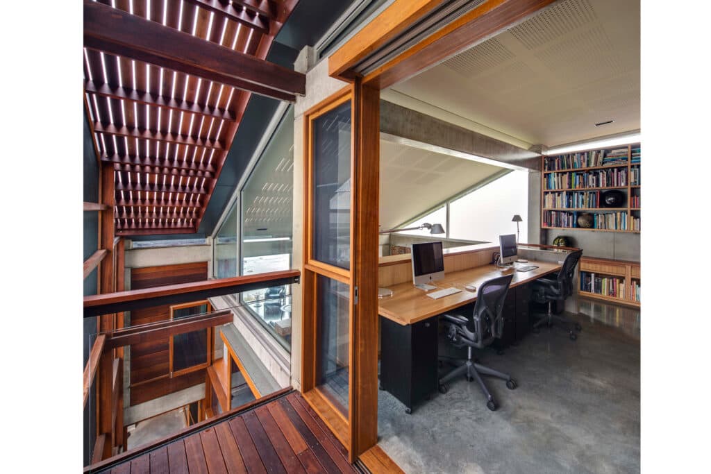 Modern office with wooden paneling, two computer desks, ergonomic chairs, and a wall of bookshelves under a slanted roof.