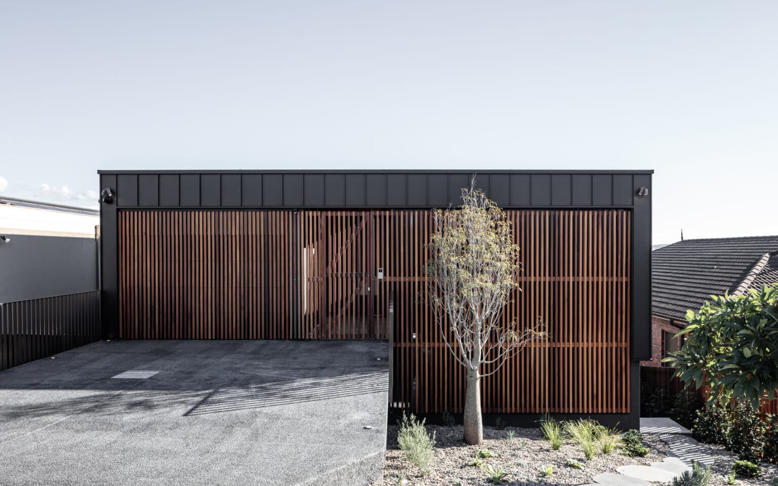 Modern black building with vertical wooden slats, a small tree in front, and a gravel driveway.