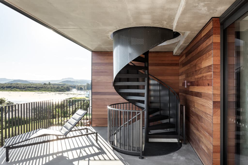 Black spiral staircase on a wooden balcony, with a lounge chair and scenic view of mountains and the beach in the distance.