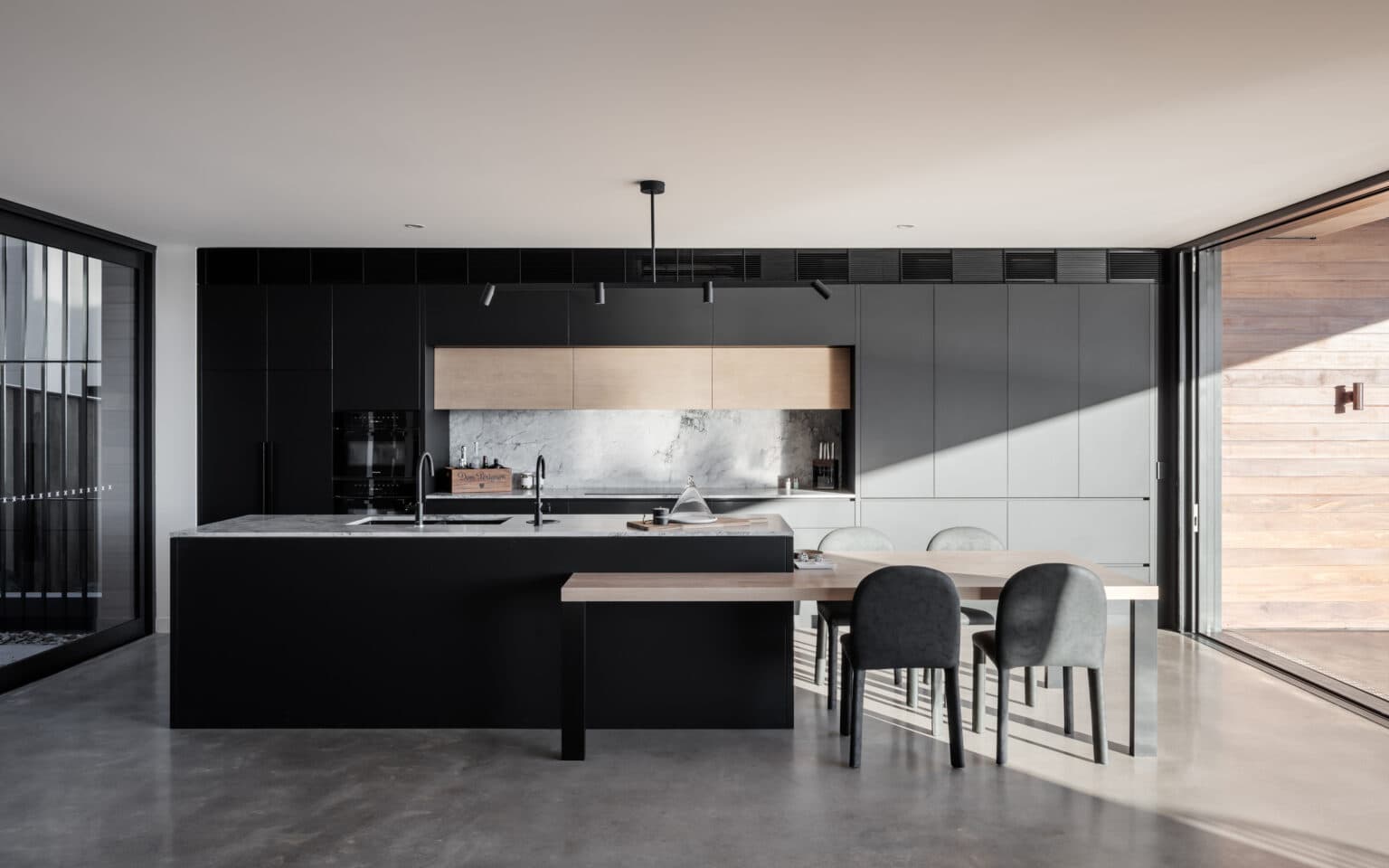 Modern kitchen with black and wood cabinets, marble countertops, island with double sink, and dining table with gray chairs.