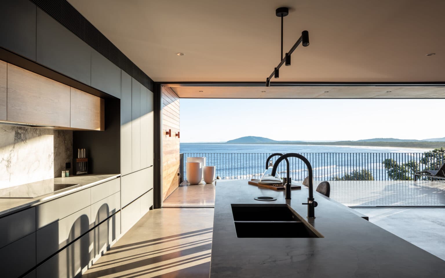 Modern kitchen with sleek cabinets opens to a balcony overlooking an ocean and mountain view, sunlit through large windows.