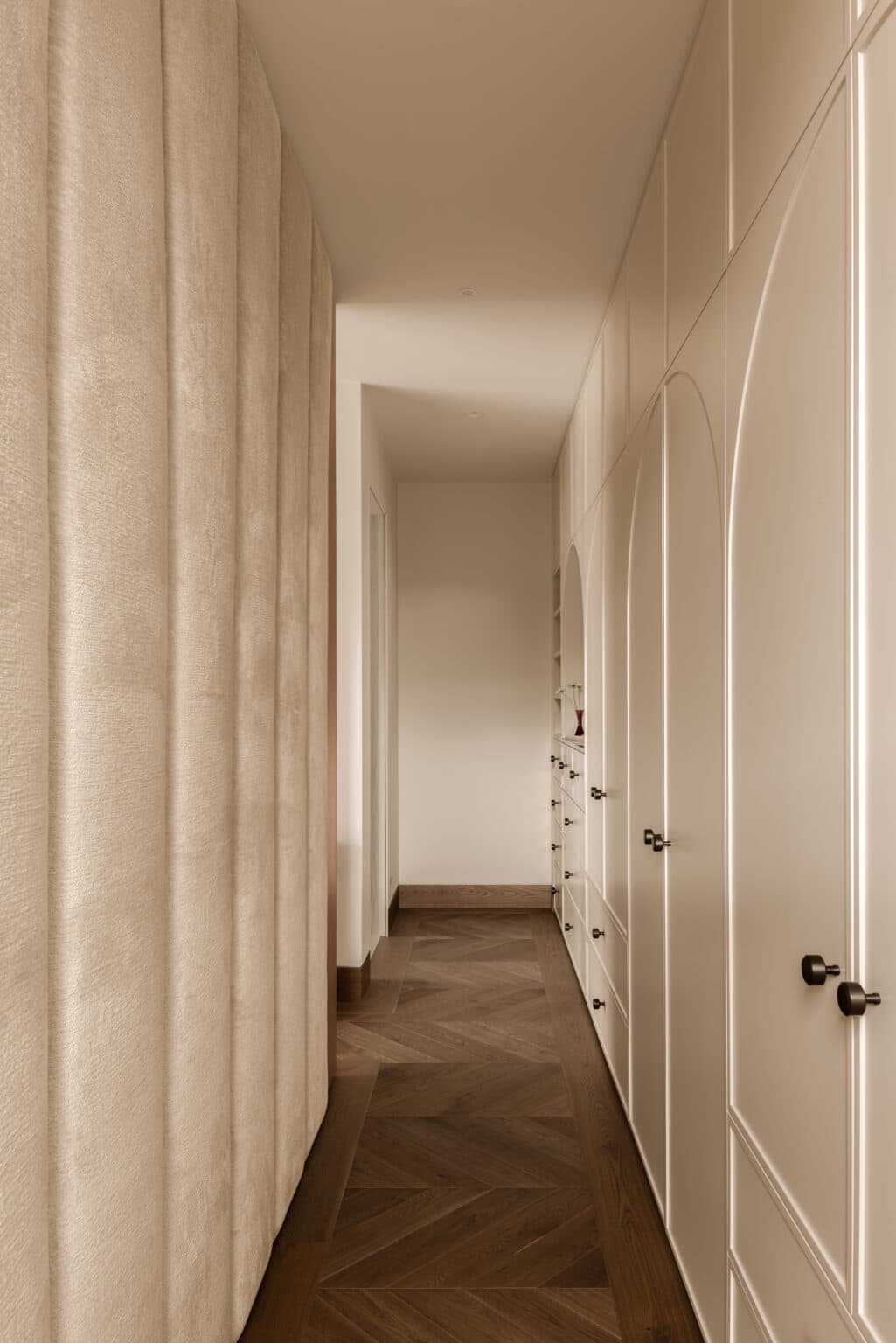 Narrow hallway with pale beige paneled walls and dark herringbone wood flooring, leading to a distant bright doorway.
