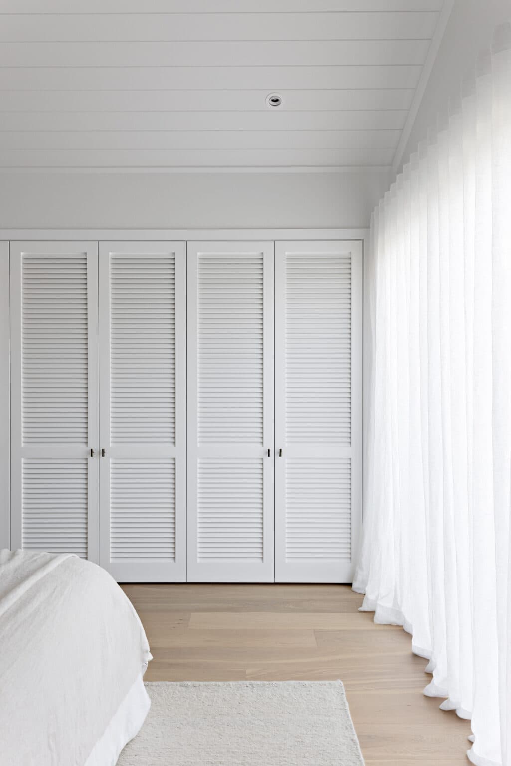 White bedroom with louvered closet doors, sheer curtains, wood floor, and a partially visible bed with a white comforter.