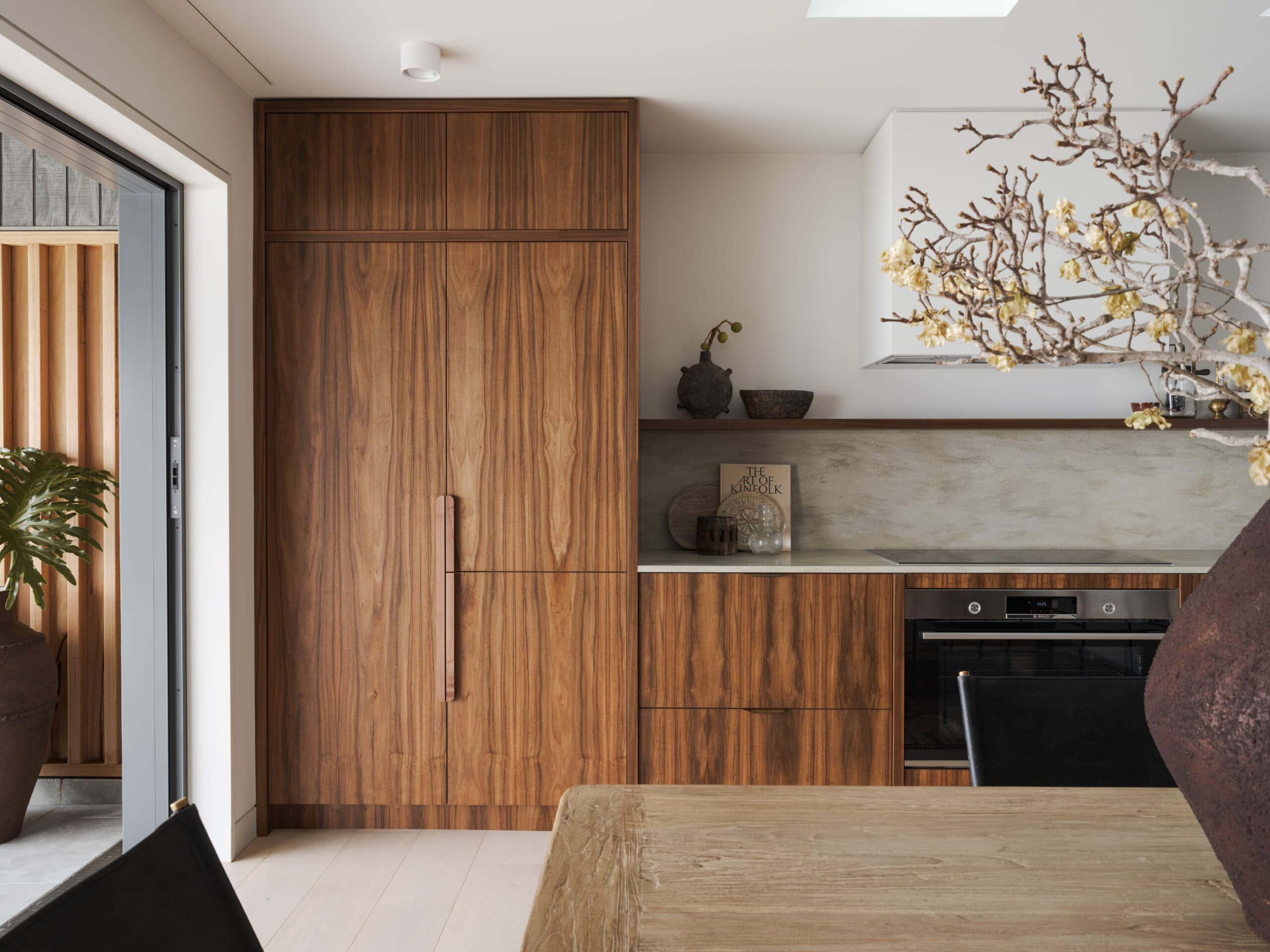 Modern kitchen with wooden cabinets, built-in oven, and a textured dining table. Decorative branches and pottery adorn the...