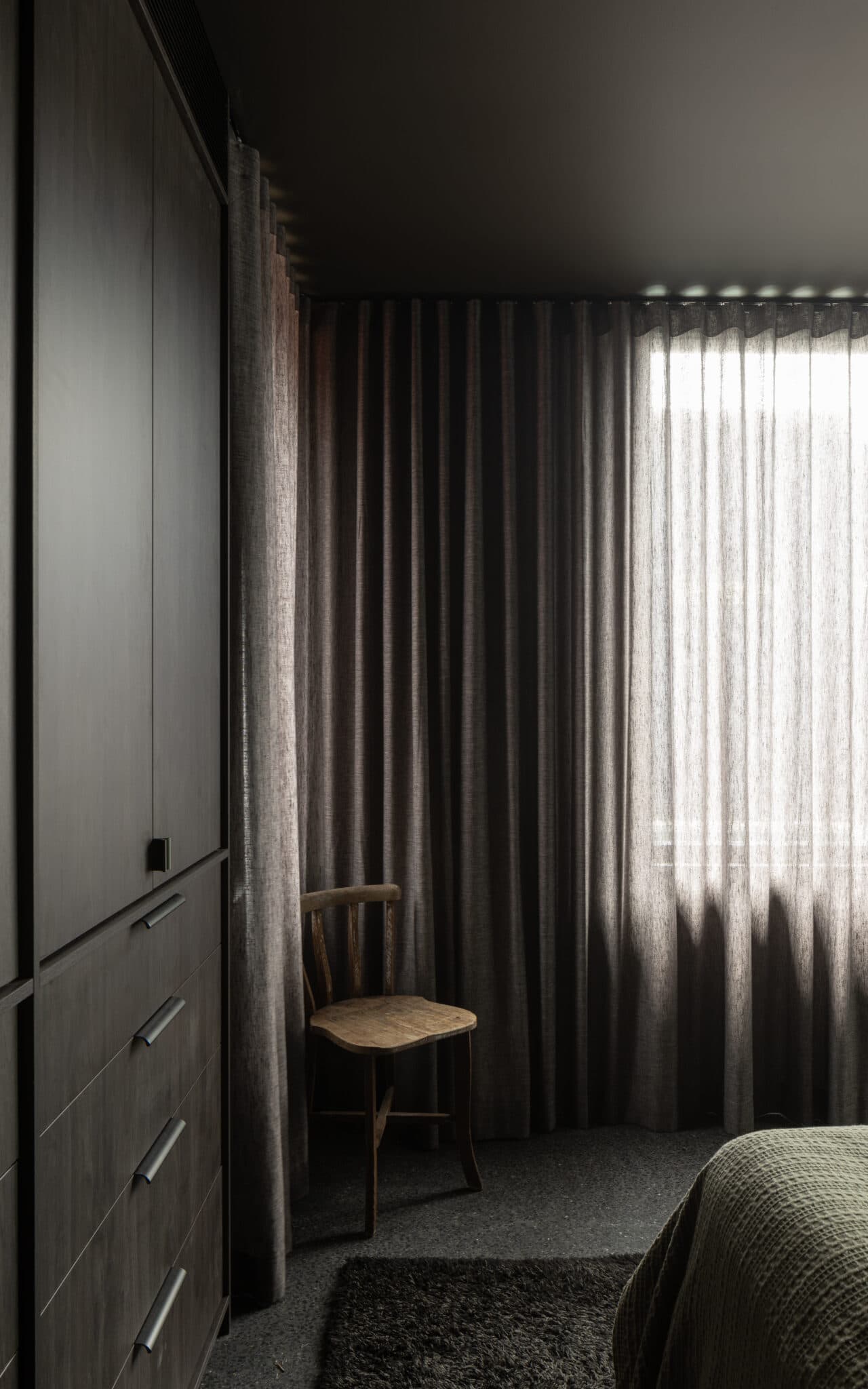 Dimly lit bedroom with dark curtains, a wooden chair, black rug, and a gray wardrobe beside a textured bed.