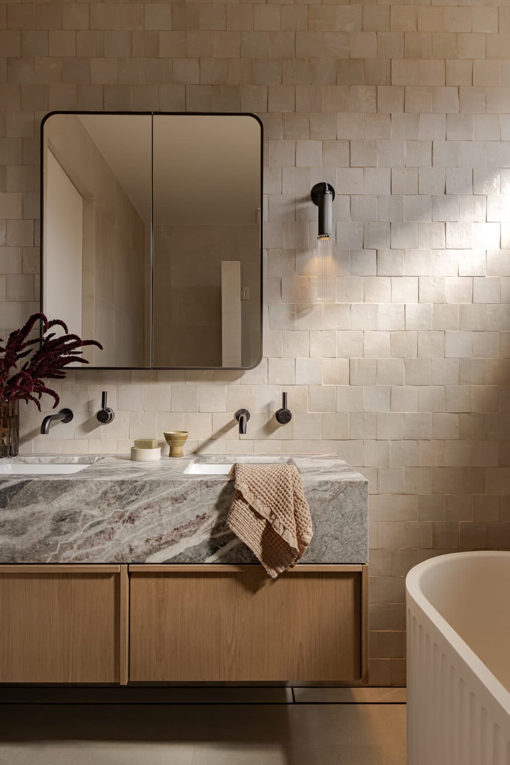 Elegant bathroom with stone countertop, dual sinks, wall-mounted faucets, beige tiles, and a hanging light fixture.