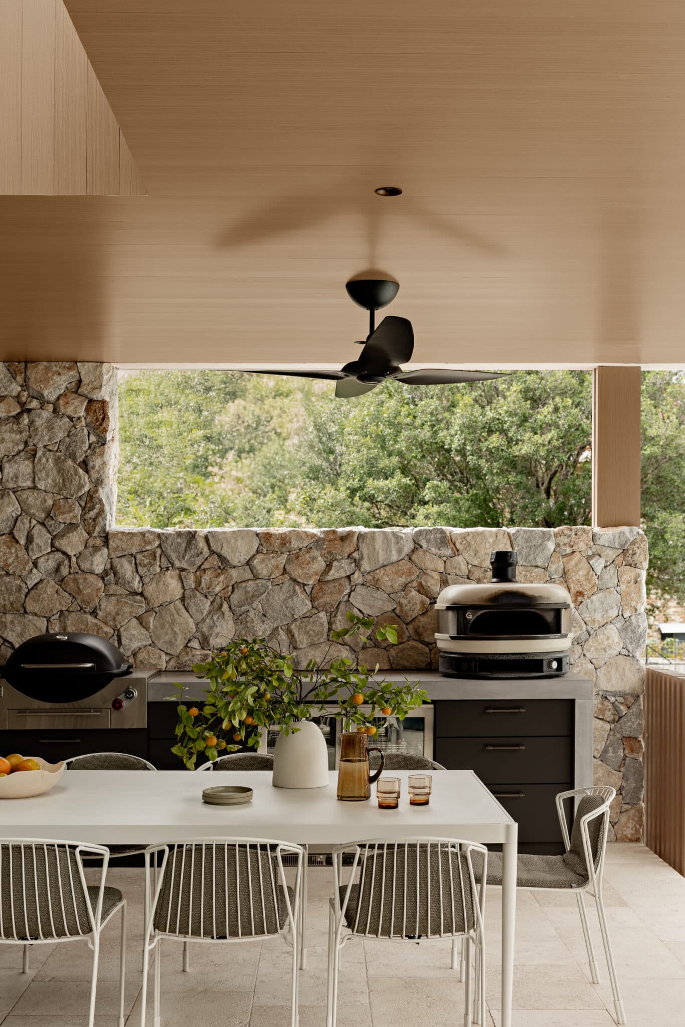 Outdoor kitchen with a stone wall, ceiling fan, white dining table, wire chairs, and a potted plant on the table.