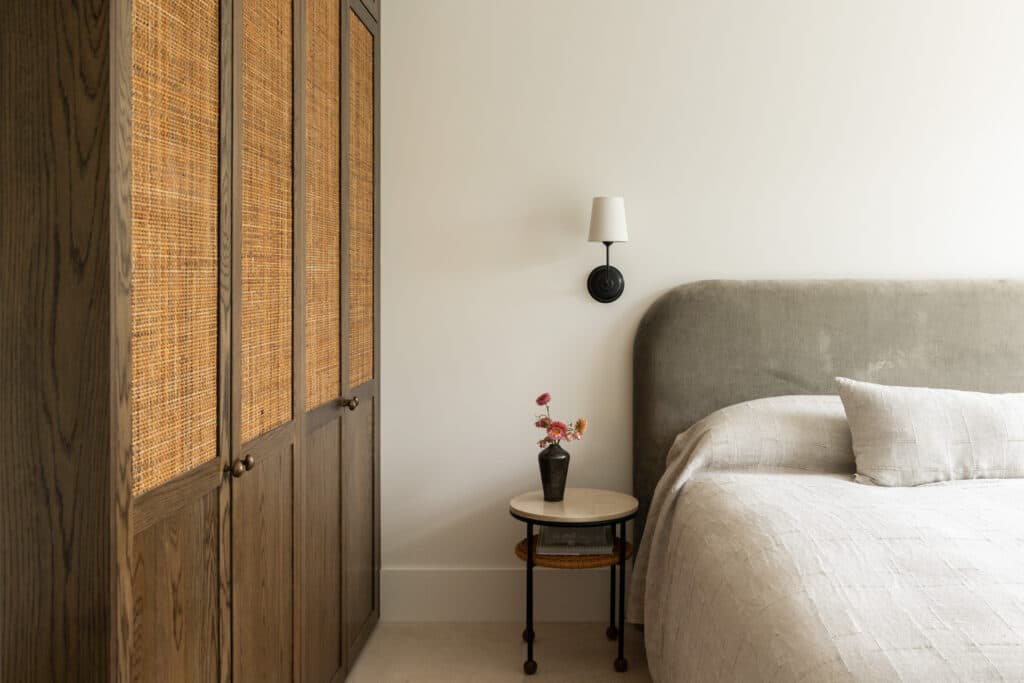 Bedroom with a gray upholstered bed, woven wood wardrobe, round bedside table with flowers, and a wall-mounted sconce.