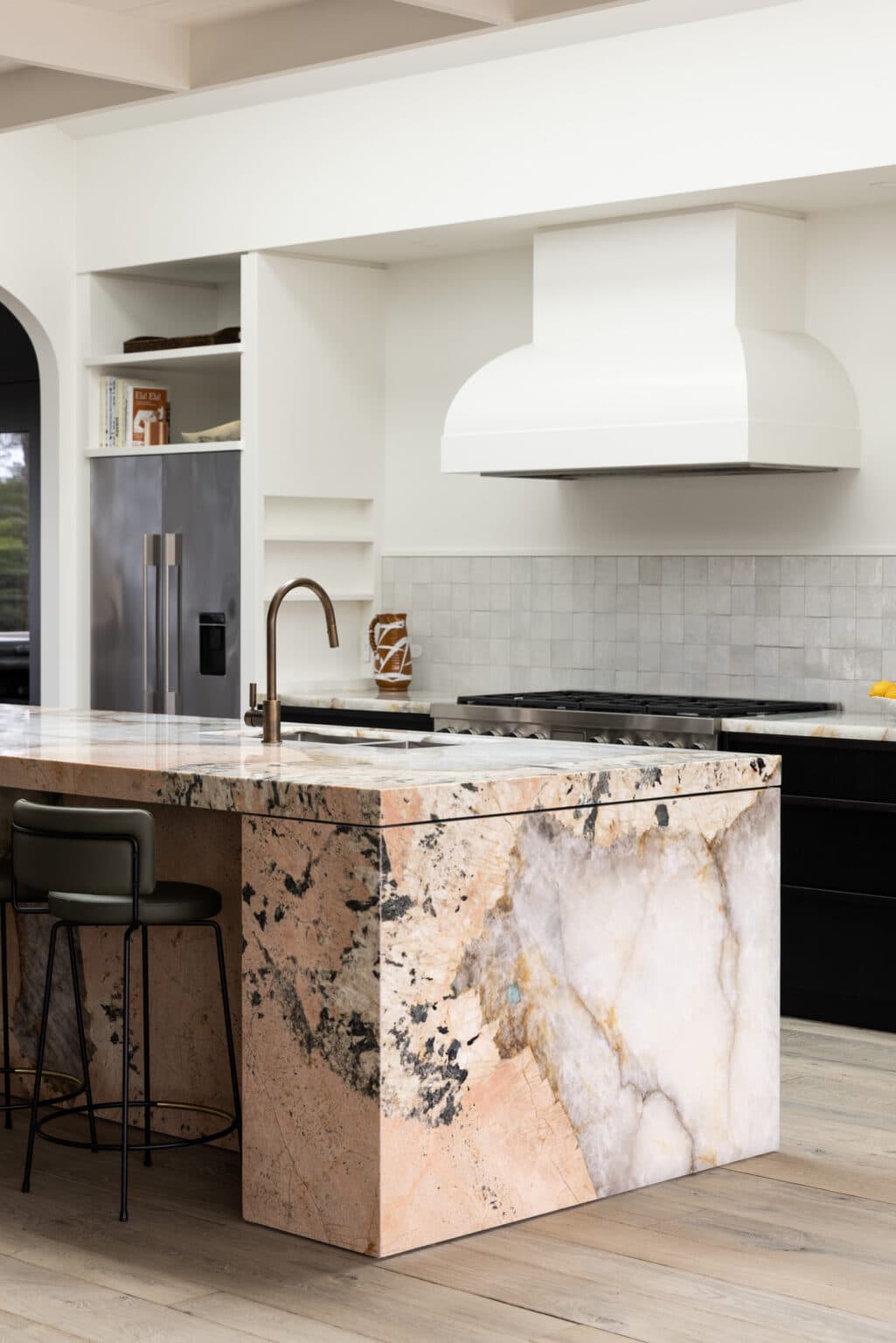 Modern kitchen with a marble island, bronze faucet, and black barstool. White cabinetry and range hood complement the look.