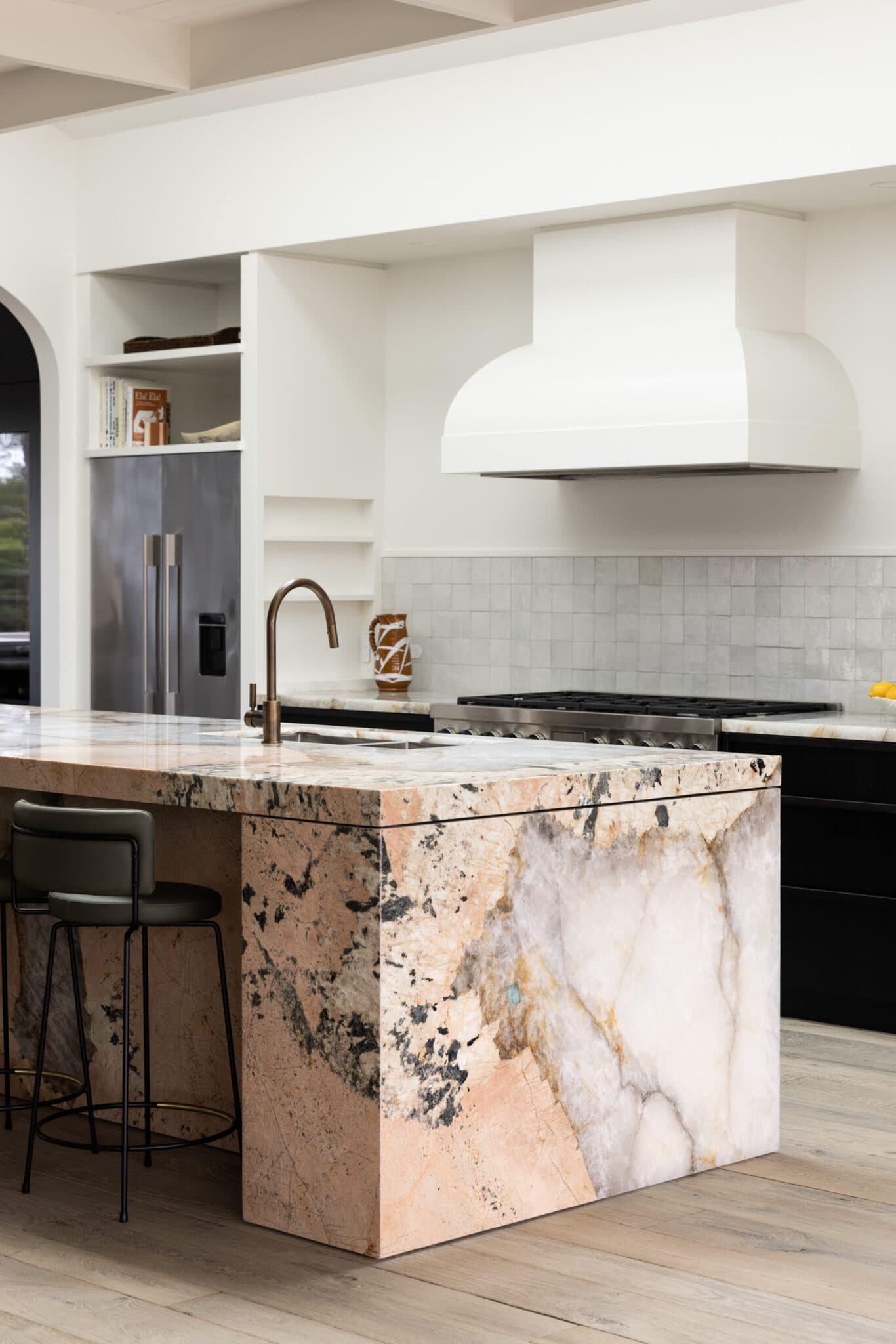 Modern kitchen with a marble island, bronze faucet, and black barstool. White cabinetry and range hood complement the look.