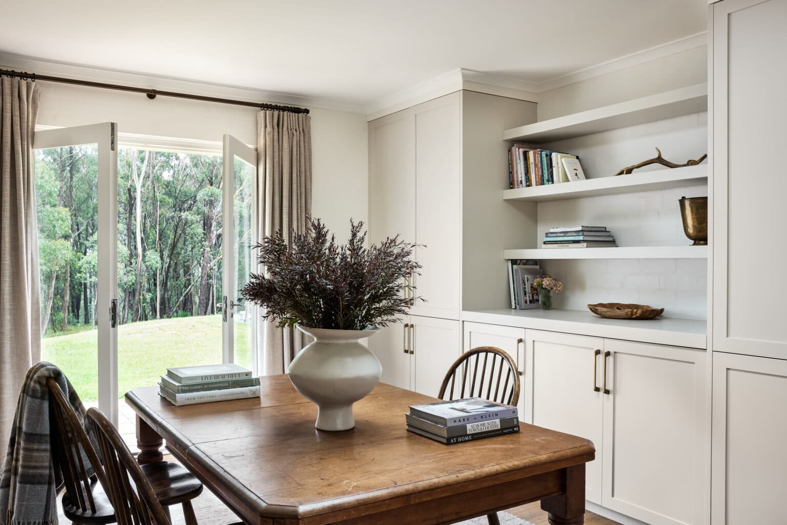 Cozy dining room with wooden table, vase of greenery, open glass doors, and built-in shelves filled with books.