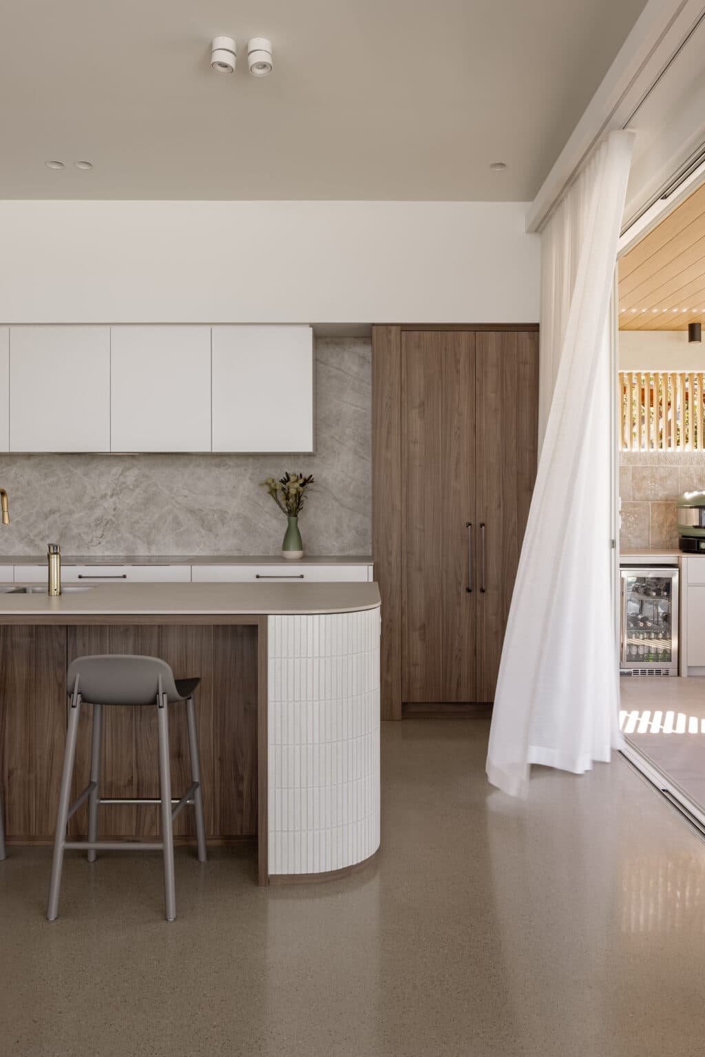 Modern kitchen with wood cabinetry, white countertops, and an open sliding door with sheer curtains leading to a patio.