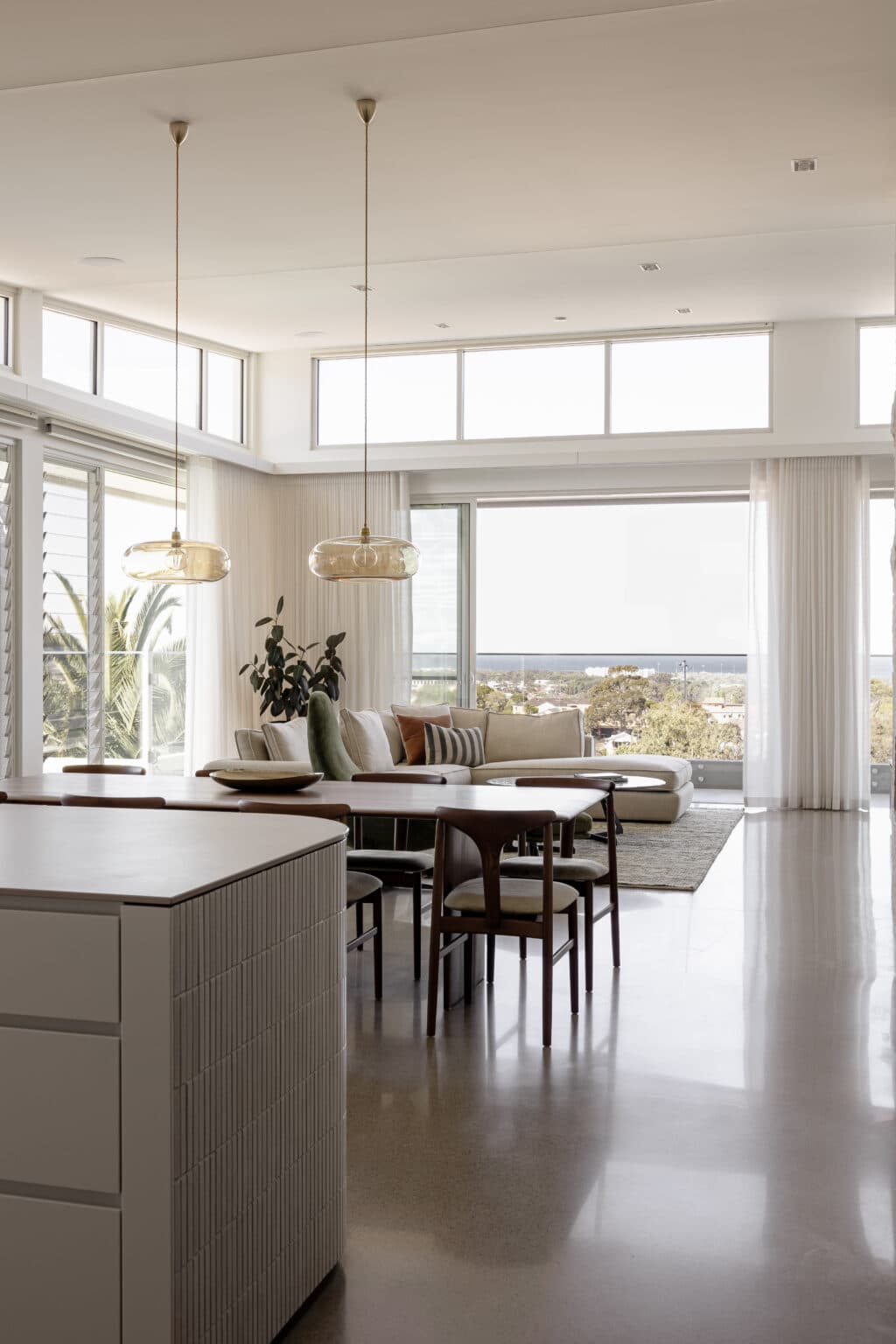 Spacious living room with large windows, round dining table, modern pendant lights, and a beige sectional sofa.
