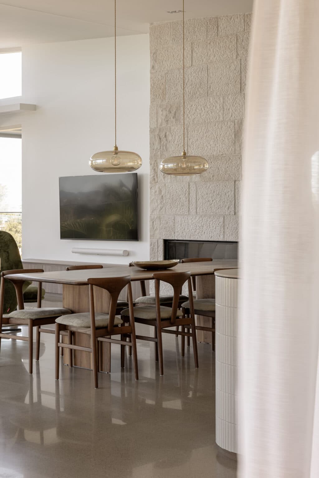 Modern dining room with wooden table, six chairs, two pendant lights, and a mounted TV on a textured white wall.