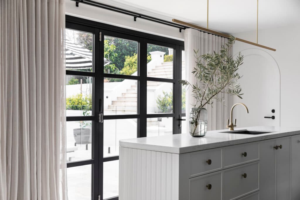 Modern kitchen with black-framed glass doors, a gold faucet, and white island counter adorned with a glass vase and foliage.