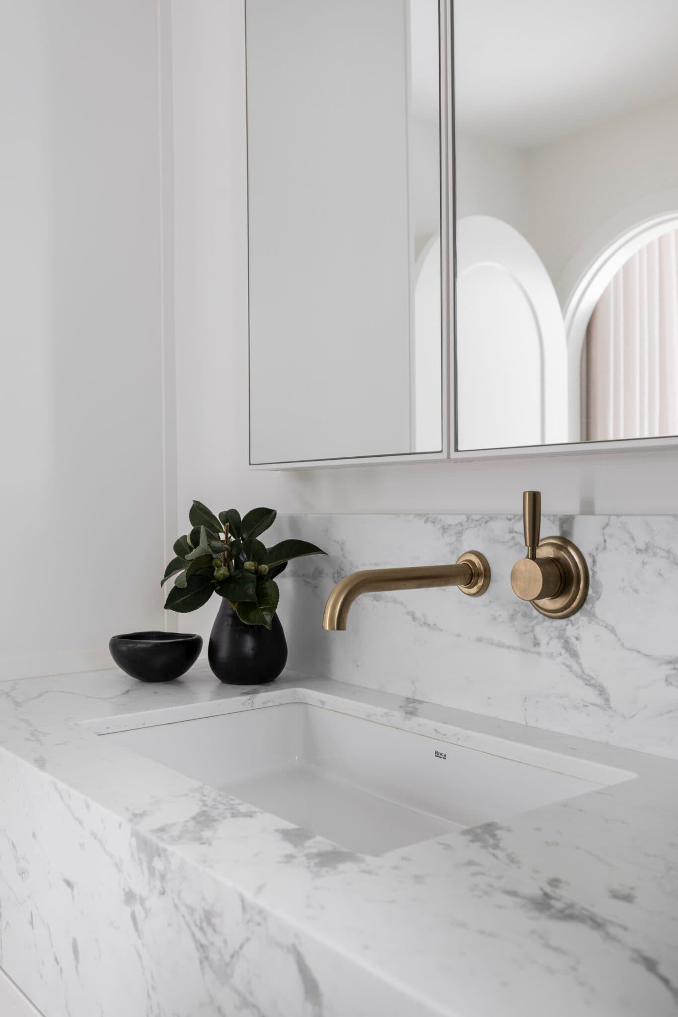 Marble sink with an elegant gold faucet, black vase with green plant, and mirror, set in a modern bathroom.