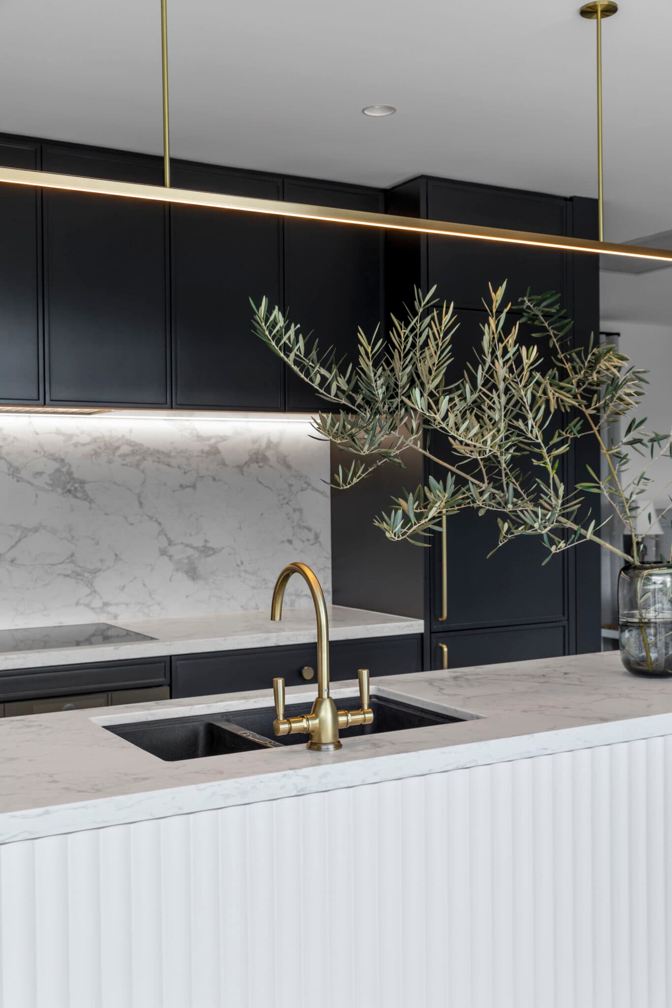 Modern kitchen with black cabinets, marble countertops, gold faucet, and a glass vase with olive branches.