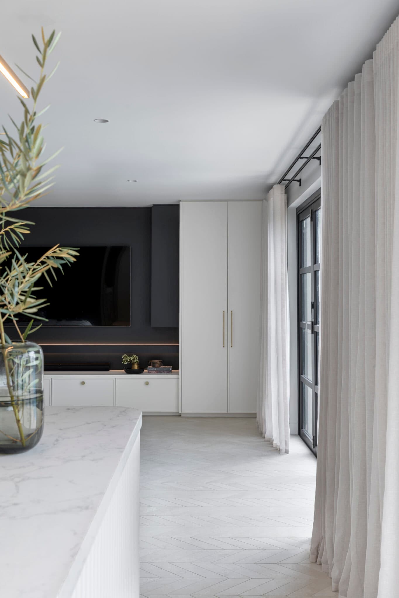 Modern living room with a marble countertop, large TV on a dark wall, white cabinets, and sheer curtains by tall windows.