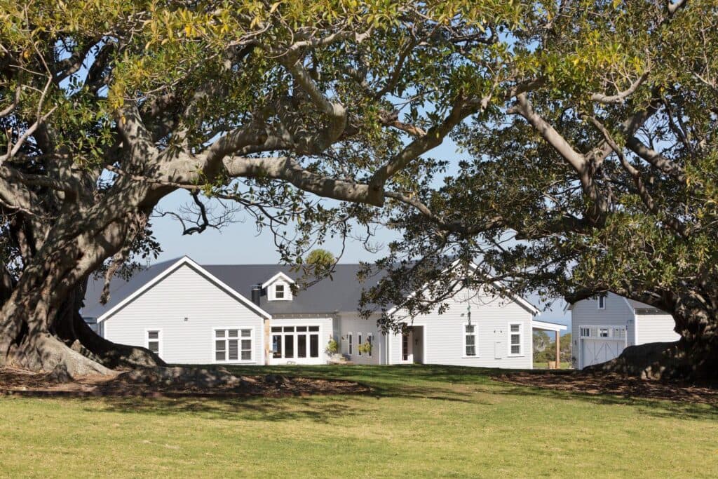 White farmhouse framed by large, leafy trees on a sunny day, with a manicured lawn in the foreground.