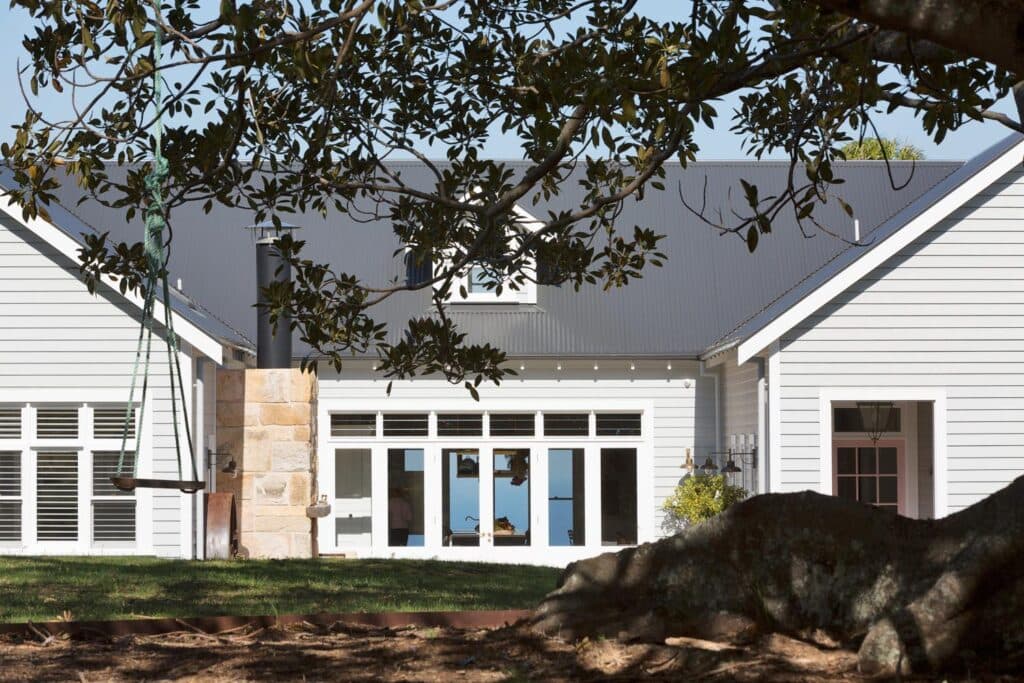 Traditional white house with large windows, a stone chimney, and a wooden swing under a leafy tree.