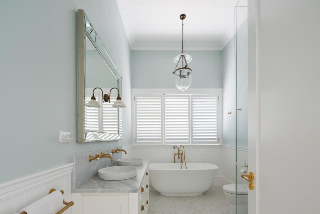 Elegant bathroom with a freestanding bathtub, double sinks, brass fixtures, and a large window with shutters.
