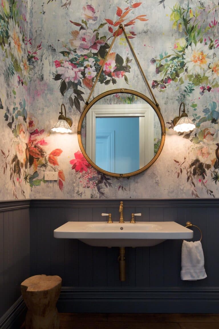 Floral wallpapered bathroom with round mirror, dual sconces, a white sink, brass fixtures, and a wooden stool.