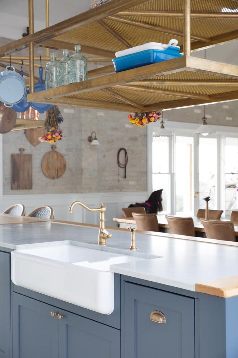 Farmhouse kitchen with blue cabinets, white apron sink, brass fixtures, hanging pots, and flowers, leading to dining area.