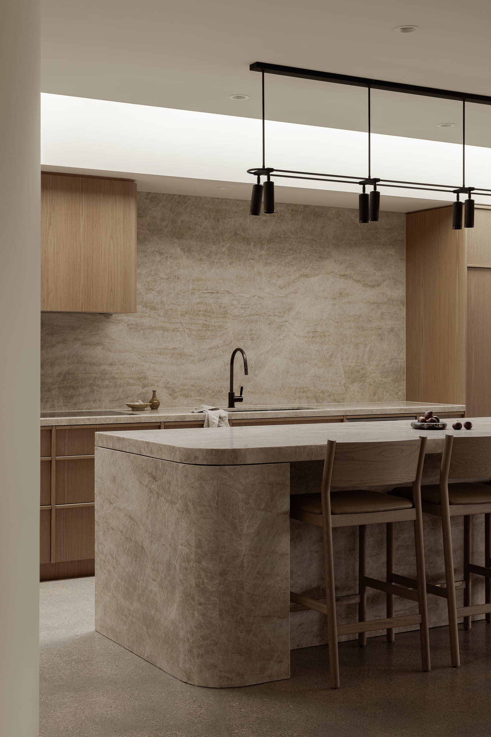 Modern kitchen with marble island, wooden cabinets, black faucet, and minimalist pendant lights. Three wooden chairs align...