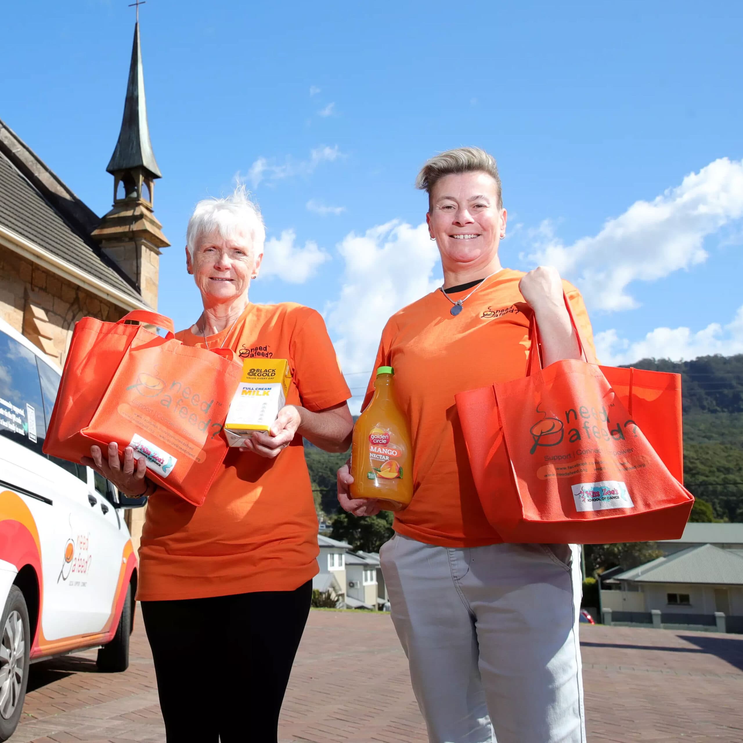 Two people in orange shirts holding grocery bags with items, standing outside a building with a pointed tower.